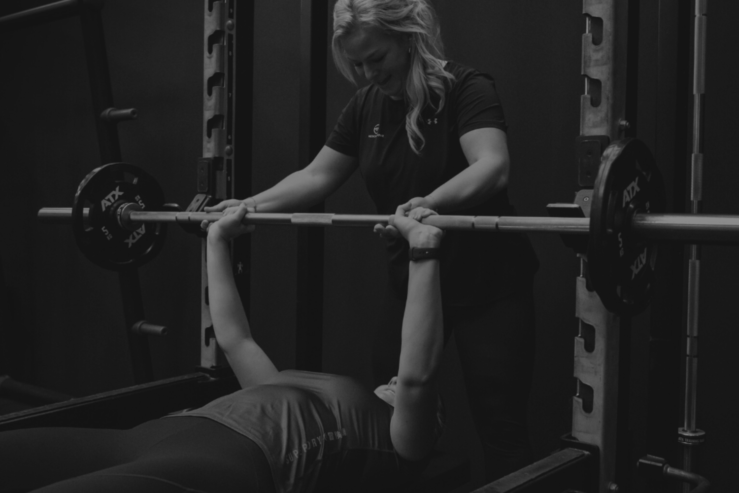 Een vrouw die onder begeleiding van een sport instructeur een benchpress uitvoert met een barbell.