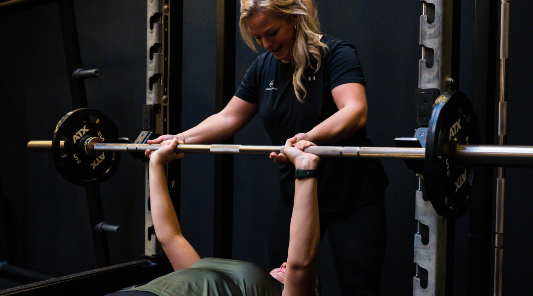 Een personal trainer die een sporter in een sportschool begeleidt bij het doen van een benchpress met een barbell waar 2 5kg gewichten aan hangen.